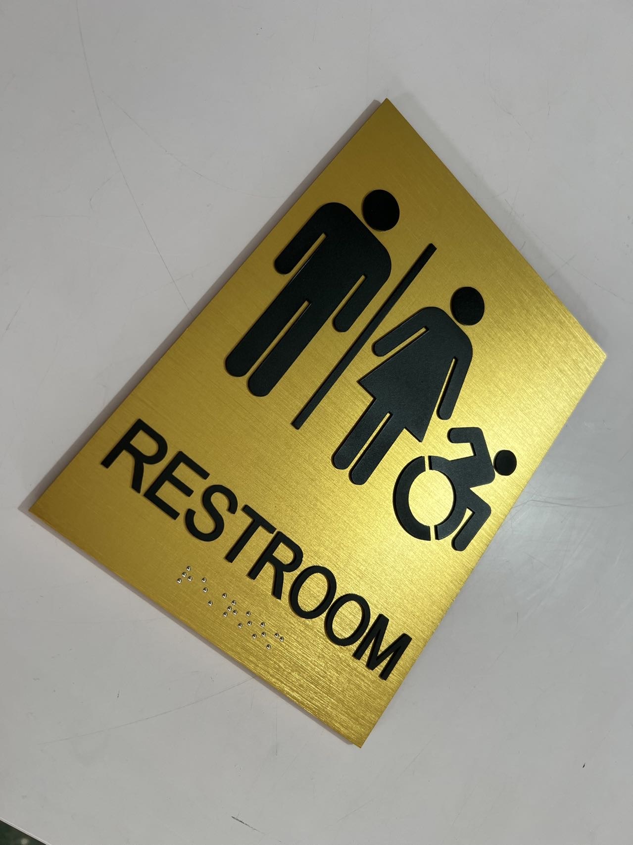 Square brushed gold accessible restroom plaque with black symbols, text and Braille